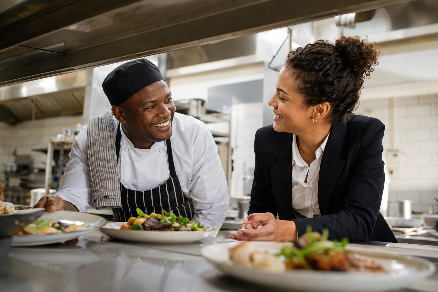 Chef and restaurant manager exchanging a smile while arranging plated dishes at kitchen pass.