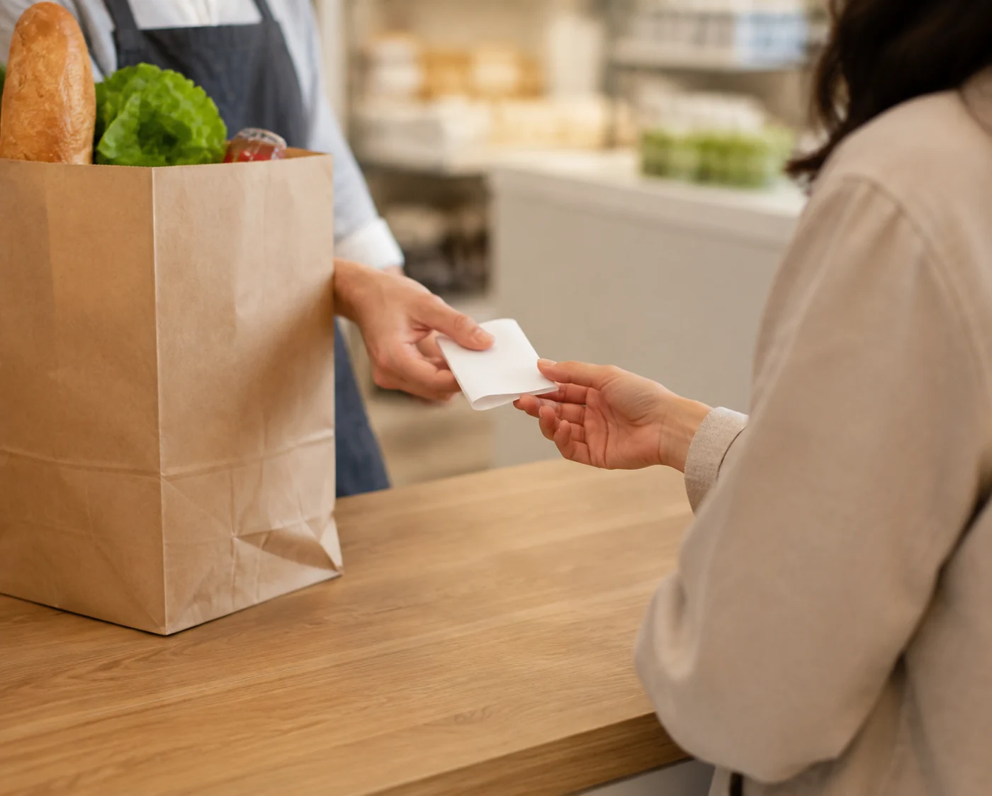 Close hands handing folded plain receipt with no readable text to customer beside packed grocery bag.